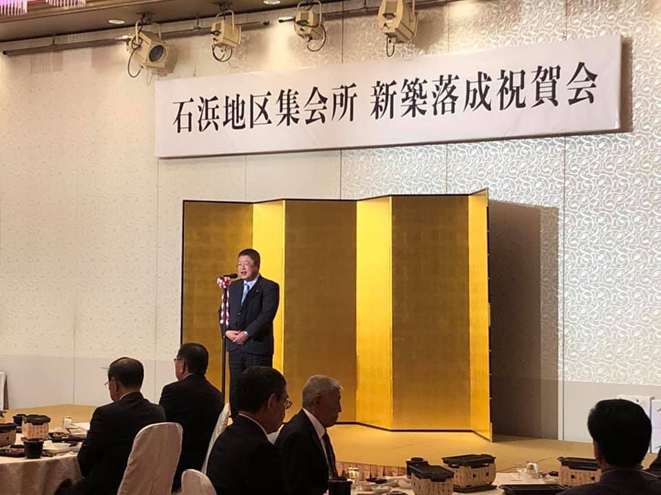 「石浜地区集会所 新築落成祝賀会」と書かれた横断幕が掲げられた壁の前に置かれた金屏風の前で、スタンドマイクに向かい話をする様子の市長の写真
