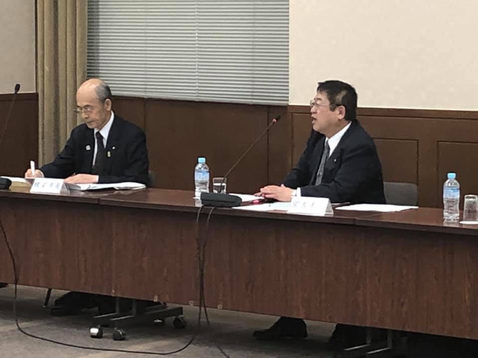 ネームプレートが置かれマイクが設置された茶色いテーブルの席に着く市長と関係者の男性を写した写真