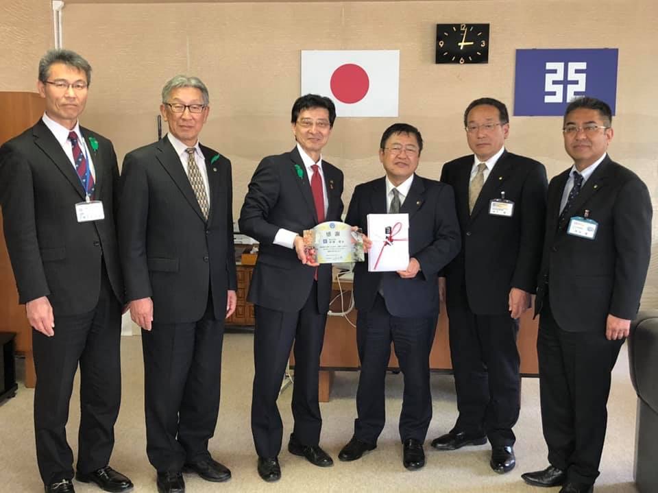 寄附金の封筒を持つ市長と、小さい盾のようなものを持つ男性を中心に、スーツを着た関係者の男性4名が横一列に並んで記念撮影をする写真