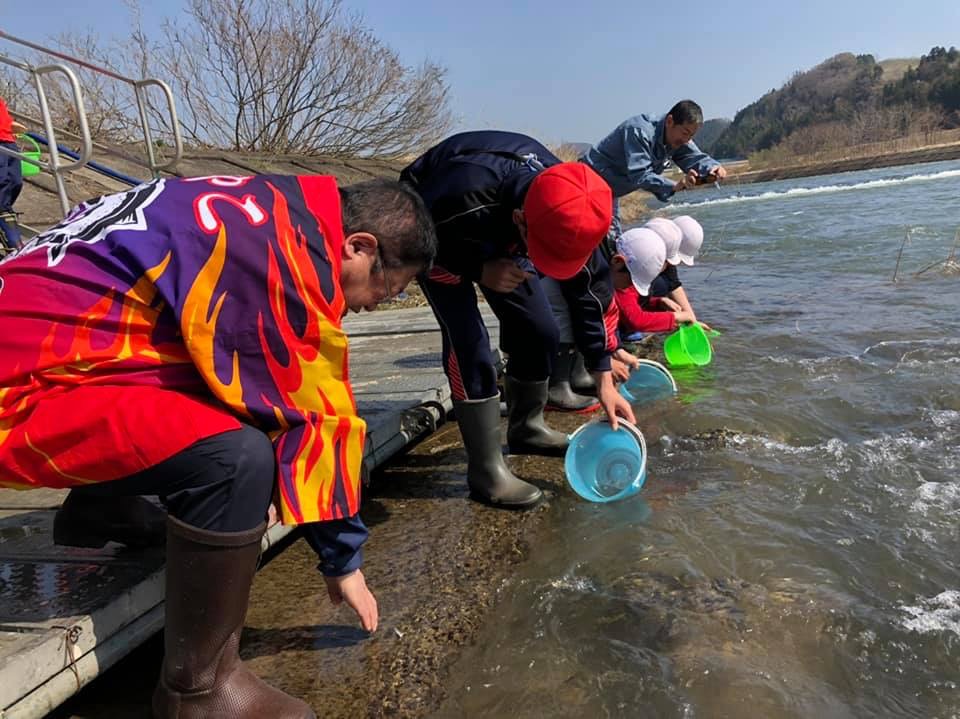 法被を着て長靴を履き、水が流れる川に右手を着けている市長と、バケツを川につけ稚魚を放流している様子の参加者たちの写真