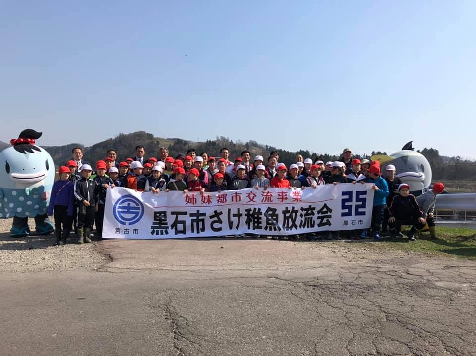 「黒石市さけ稚魚放流会」と書かれた横断幕を前列で持つ小学生たちと、その後ろに並ぶ小学生と関係者の方々、両側に立つサーモンくんとみやこちゃんの記念写真