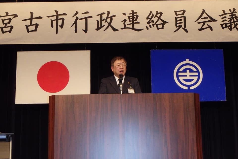 「宮古市行政連絡員会議」と書かれた横断幕、日本国旗、宮古市の市町村旗が掲げられた舞台で、演台に置かれたマイクに向かい話をする様子の市長の写真