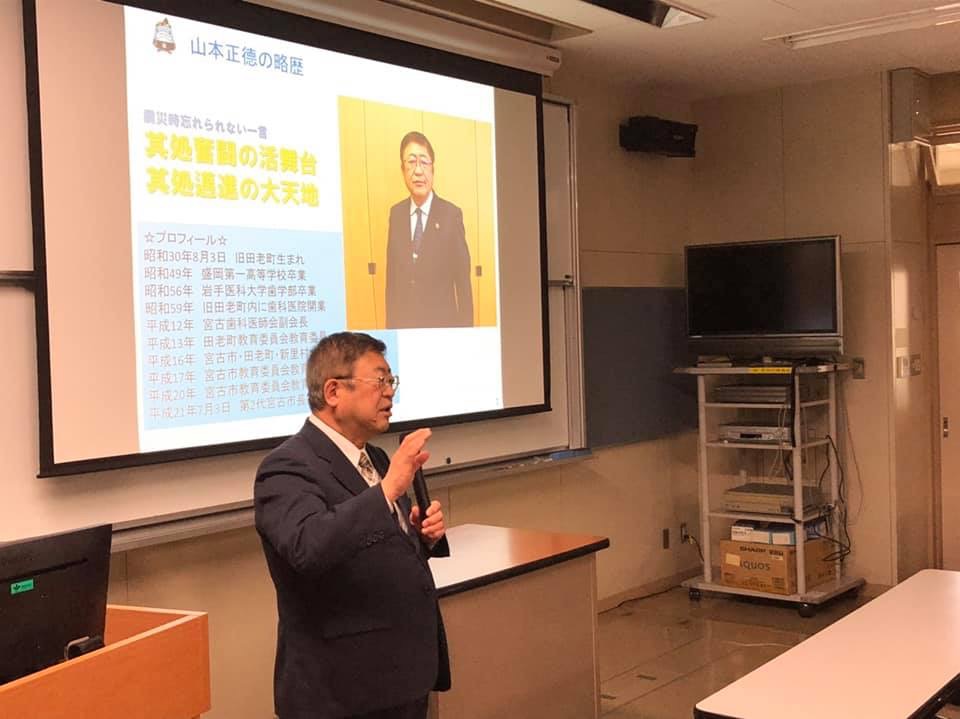 市長の写真が投影されたスクリーンの斜め前方に立ち、左手にマイクを持って講演する様子の市長の写真