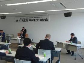 壁に「第17期定時株主総会」と書かれた横断幕の下の演台の前で発表されているスーツ姿の関係者と参加者が話を聞いている写真