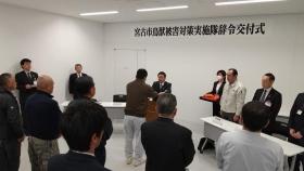 「宮古市鳥獣被害対策実施隊辞令交付式」と書かれた紙が離れた会場で、任命書を渡す市長と受け取る様子の男性、その周りに立つ関係者の方々の写真