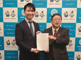 バックパネルの前で任命書を一緒に持つ様子の市長と男性の記念写真