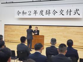 「令和2年度辞令交付式」と書かれた横断幕が吊るされた会場で、演台で市長から辞令を受け取る男性、その後方で椅子に座って辞令交付を待つ職員の方々の写真