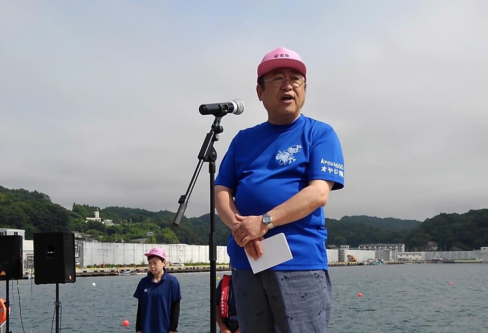 後方に海が見える宮古市魚市場前で、ピンク色のキャップに青色のシャツを着た市長がマイクスタンドの前で挨拶を行っている様子の写真