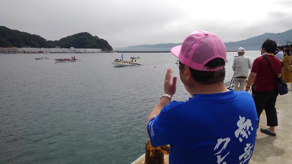宮古港の海上で行われているカッターレースを、観客やピンク色のキャップに青色のシャツを着た市長が応援している後ろ姿の写真