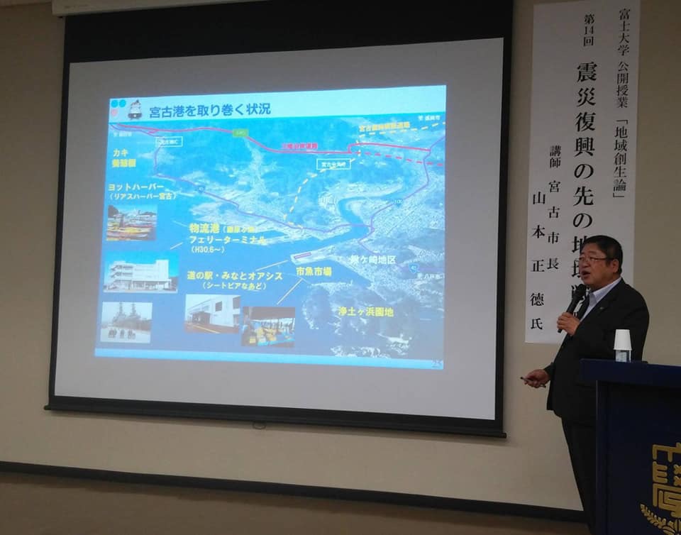 資料が映し出さされたスクリーンの右側に「第14回震災復興の先の地域戦略」と書かれた幕が掲げられ、手前に立つ市長が左手にマイクを持ち話をしている講義の様子の写真