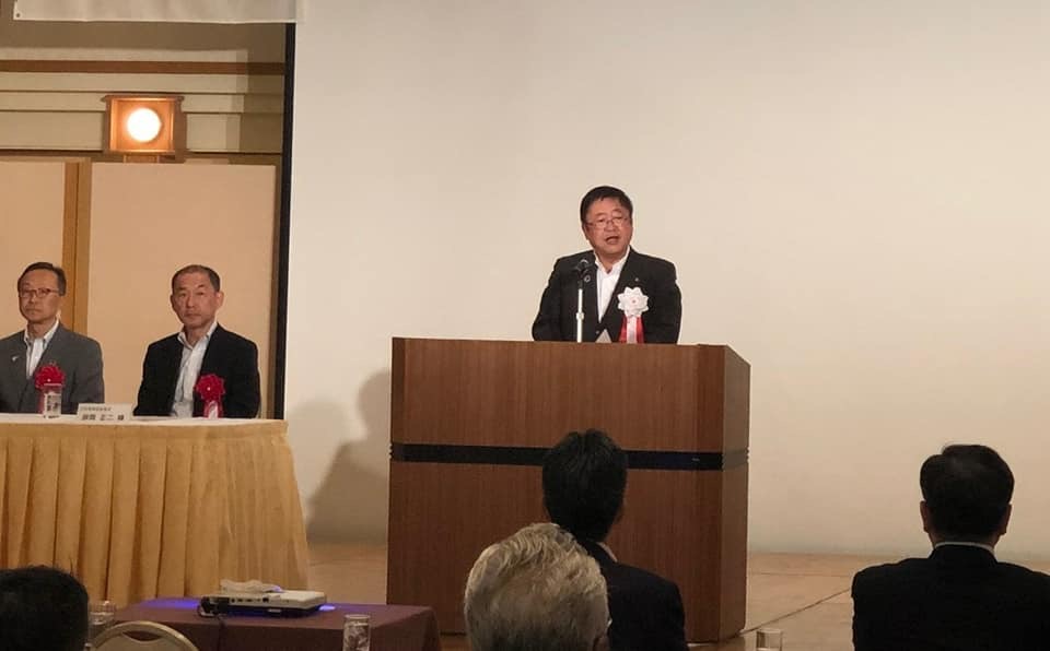 着席した関係者の方々を前に、左胸に胸章リボンを付けた市長が檀上で挨拶を行っている総会の様子の写真