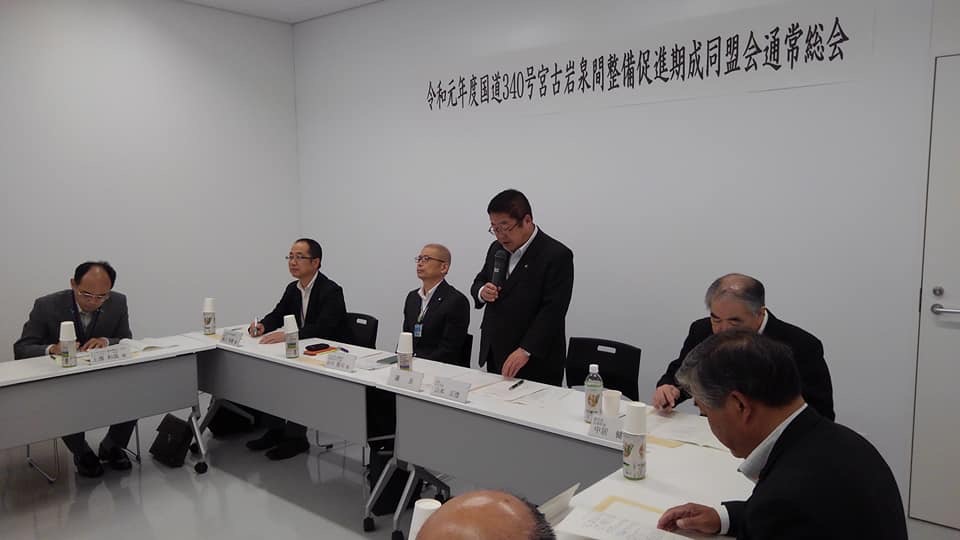「令和元年度国道340号宮古岩泉間整備促進期成同盟会総会」と書かれた横幕が壁に貼られた室内の形に合わせて長机が設置された席に関係者が座り、中央の席から立ち上がった市長が右手でマイクを持ち挨拶を行っている総会の様子の写真