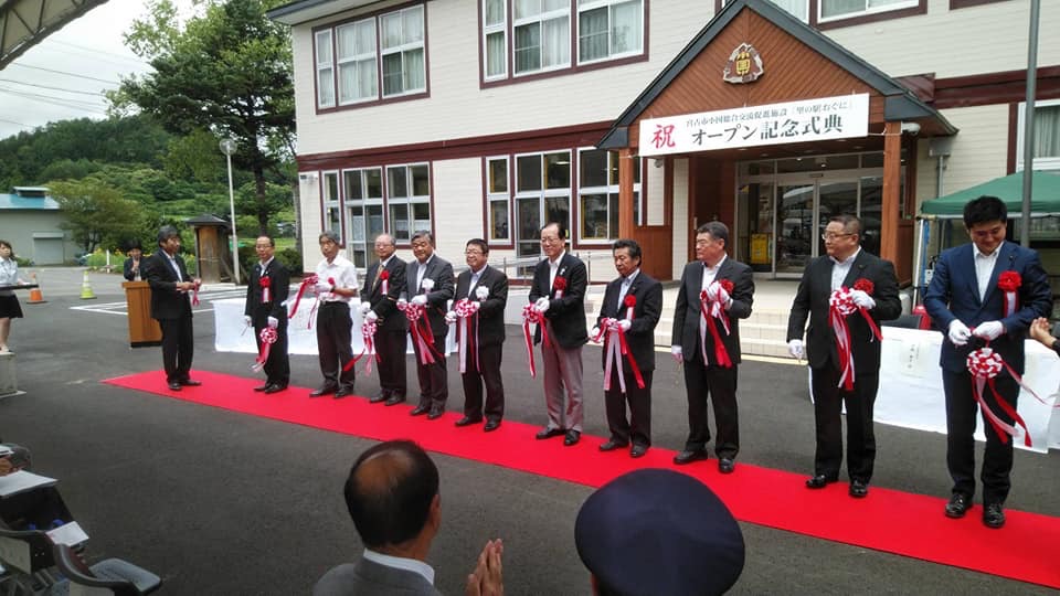 建物の入り口の上に「祝オープン記念式典」と書かれた横断幕が貼られ、レッドカーペットが敷かれた屋外で、市長を中央に10名の関係者が横一列に並びテープカットが行われた様子の写真
