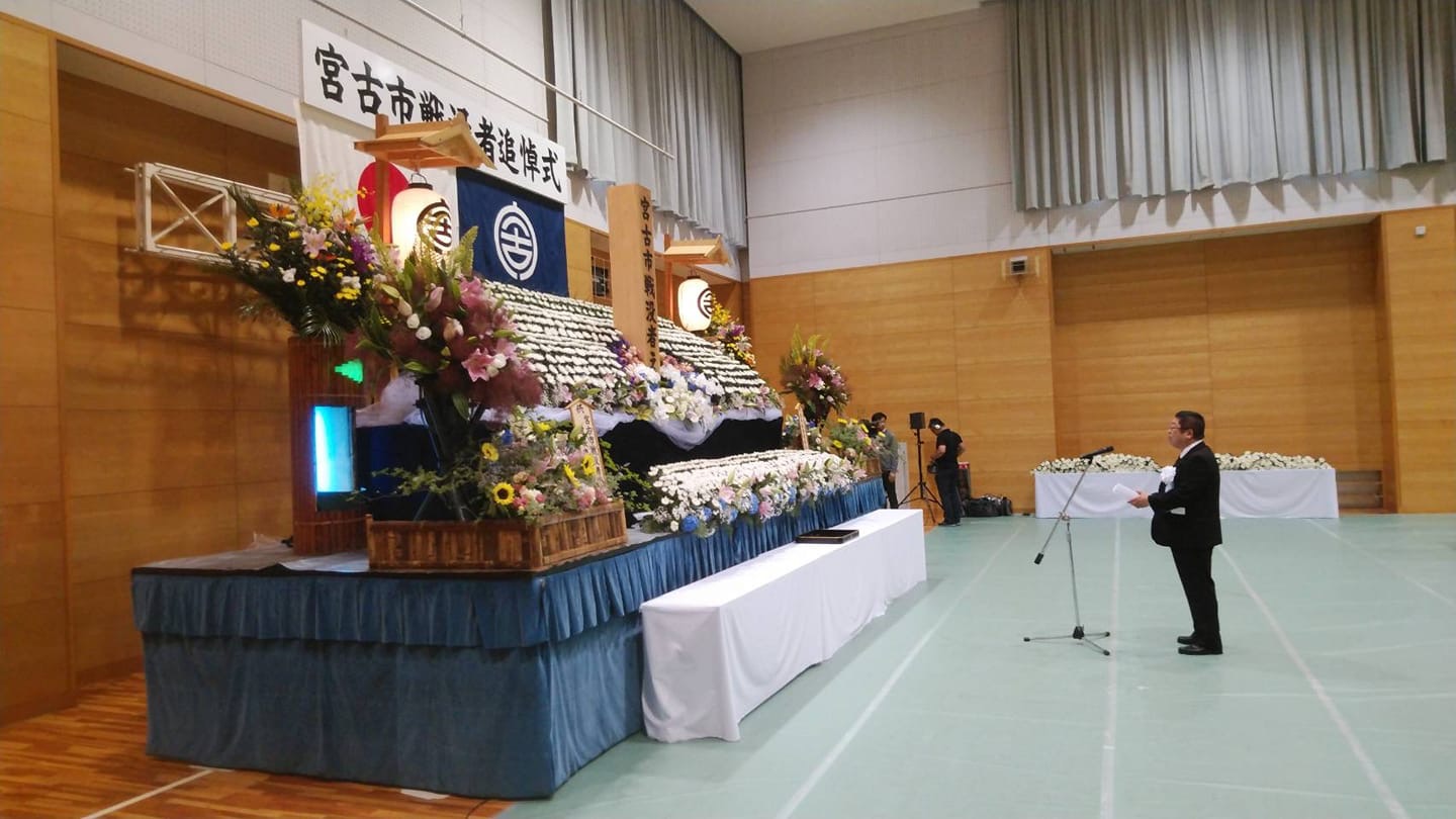 前方に大きく書かれた「宮古市戦没者追悼式」の横幕に国旗と市旗が掲げられ、沢山の花で飾られた祭壇前のマイクスタンドの傍で市長が挨拶を行っている様子を左側から撮影した写真
