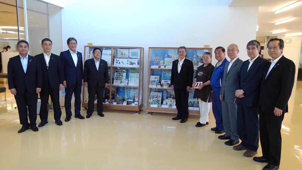 特産品が展示された背丈くらいある大きさの2つのショーケースが展示され、左右に市長や副市長、8名の関係者が扇形に並んでいる写真