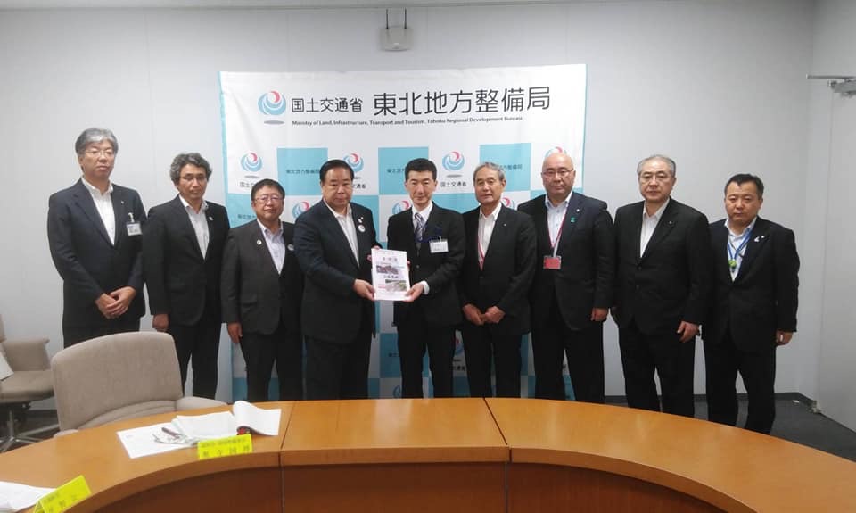 国土交通省東北地方整備局と書かれたバックパネルの前で、中央に東北地方整備局の佐藤克英局長と同盟会の代表が一緒に要望書を持ち、市長を含む7名の関係者が横一列に並んでいる写真