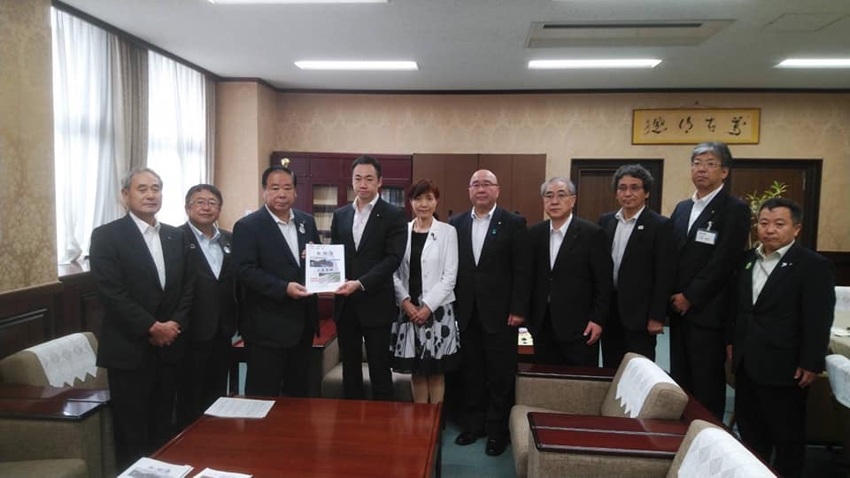 応接用のテーブルや椅子が設置された室内で、中央に財務省の鈴木馨祐副大臣と同盟会の代表が一緒に要望書を持って中央に立ち、市長や高橋ひな子衆議院議員を含む8名の関係者が横一列に並んでいる写真
