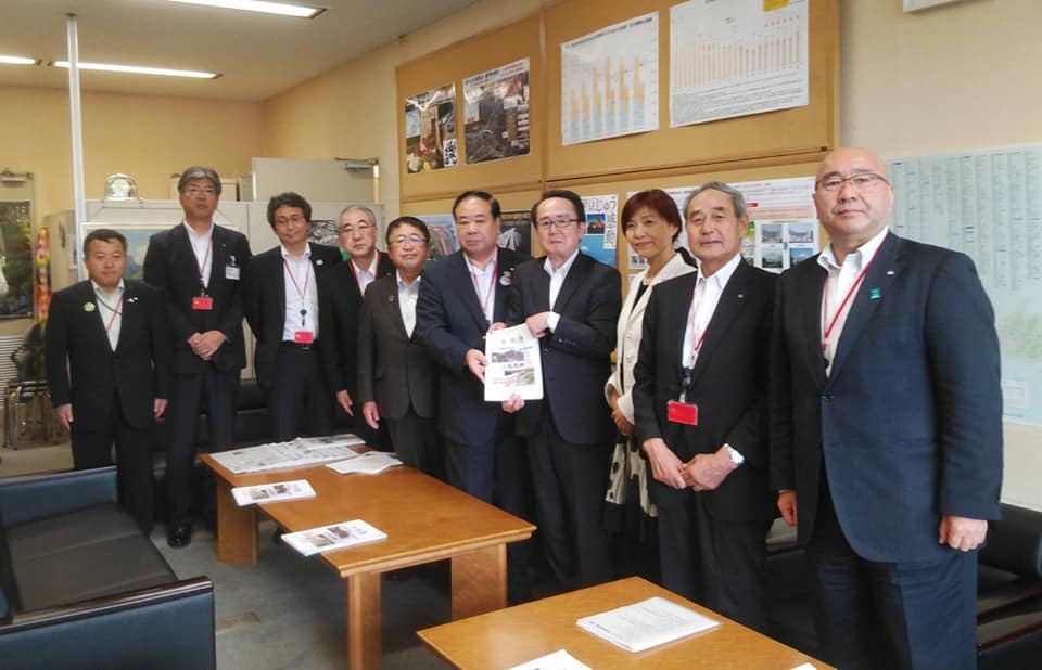壁に大きなポスターやグラフが掲示された室内で、中央に国土交通省の池田豊人道路局長と同盟会の代表が一緒に要望書を持って中央に立ち、市長や高橋ひな子衆議院議員を含む8名の関係者が横一列に並んでいる写真
