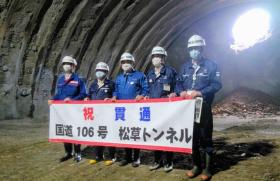 工事中のトンネルの中で、「祝貫通 国道106号 松草トンネル」と書かれた横断幕を持つ、ヘルメットを被りマスクを着けた5名の関係者の方々の記念写真