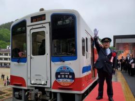青、白、赤のカラーリングがされた電車の横で、制帽を被り白い手袋を着けて右手を上に大きく上げている様子の市長の写真