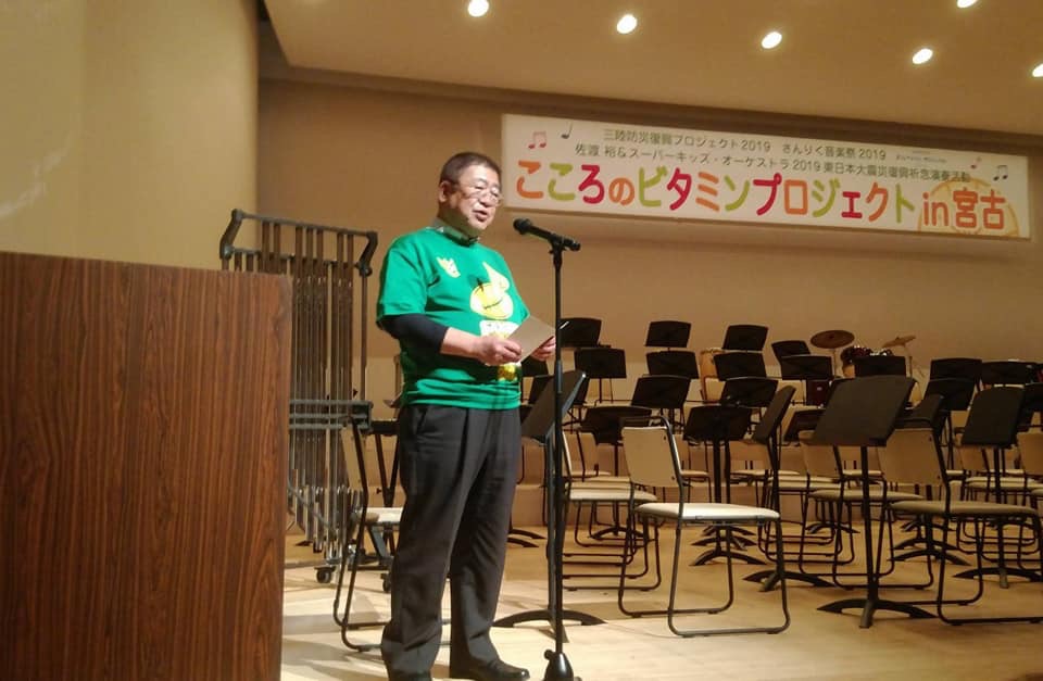 「こころのビタミンプロジェクトin宮古」と書かれた横断幕が吊るされ、椅子や譜面台が並べられた会場で、スタンドマイクに向かい話をする市長の写真