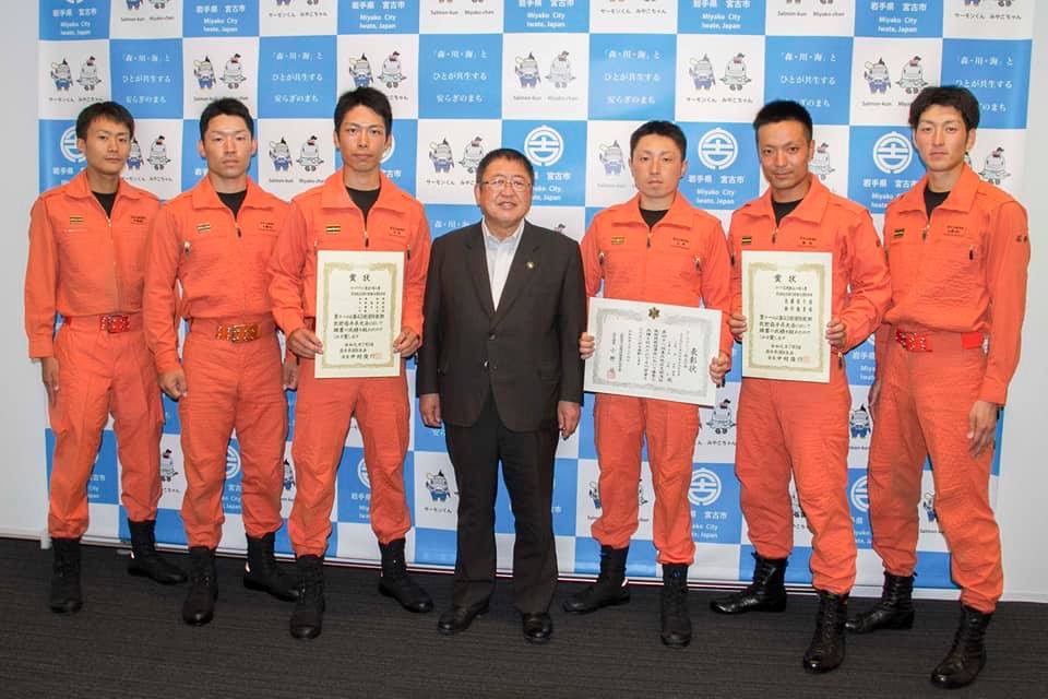 バックパネルの前で、市長を中心に並ぶオレンジ色の隊服を来た宮古地区広域行政組合の救助隊員6名の内3名が賞状を持っている記念写真