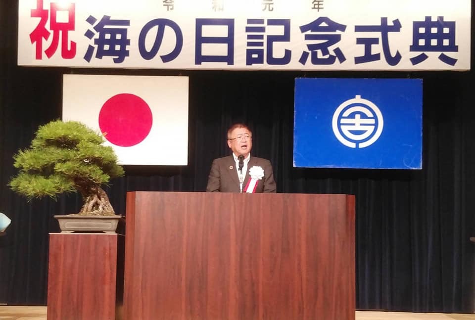 2枚の旗と「令和元年 祝 海の日記念式典」と書かれた横断幕が掲げられた檀上で、左胸に胸章リボンを付けた市長が挨拶を行っている様子の写真
