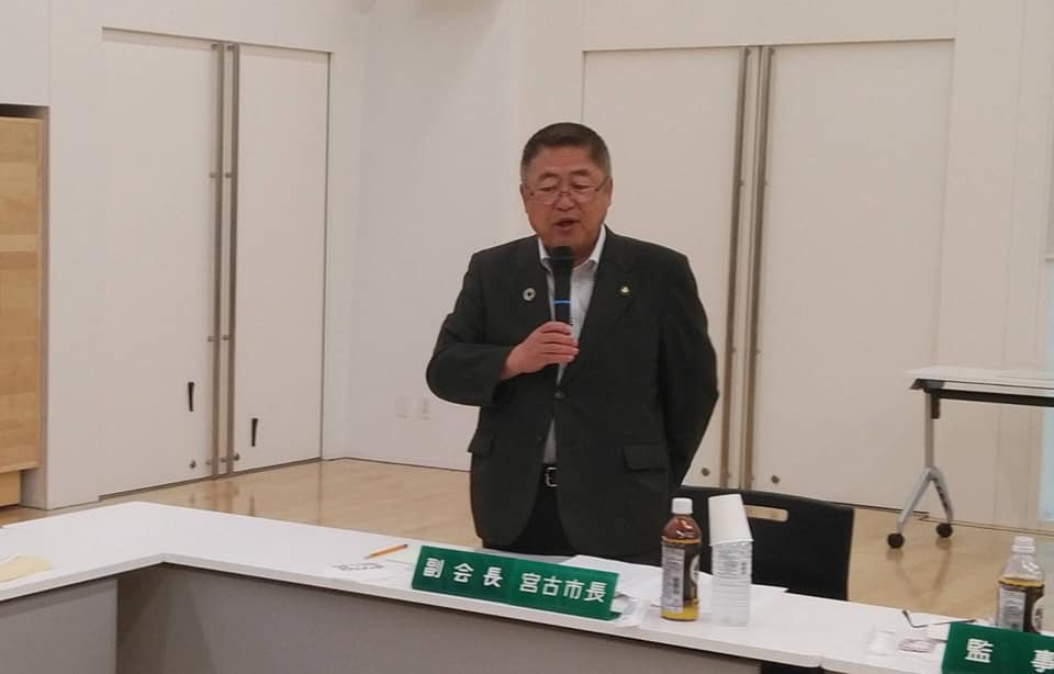 「副会長 宮古市長」と書かれた席札が置かれた席から立ち上がった市長が、右手にマイクを持ち話をしている写真