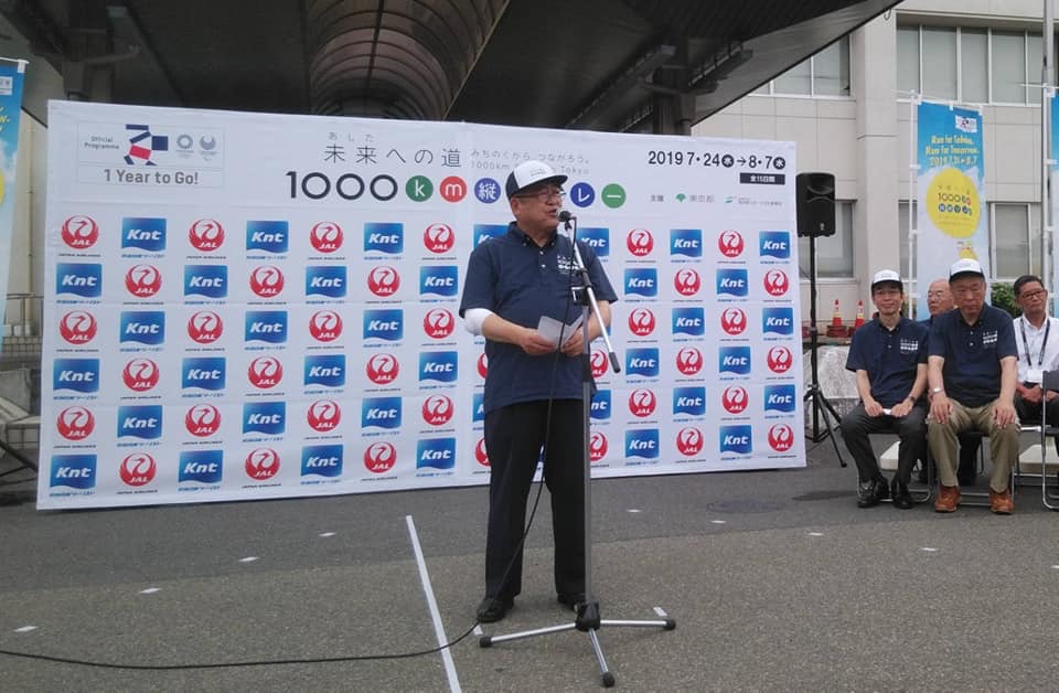 合同庁舎建物入り口に設置された「未来（あした）への道 1000キロメートル縦断リレー 2019」のバックパネルの前で、キャップを被った市長がマイクスタンドの前で挨拶を行っている様子の写真
