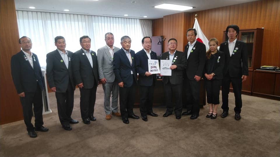 国旗が飾られた室内で、中央に渡辺博道復興大臣と市長が一緒に要望書を持ち、8名の関係者が横一列に並んでいる写真