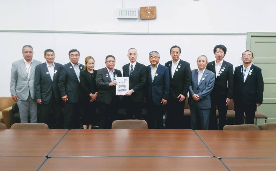 白い壁の上に時計が飾られた室内で、中央に財務省の太田充主計局長と市長が一緒に要望書を持ち、9名の関係者が横一列に並んでいる写真