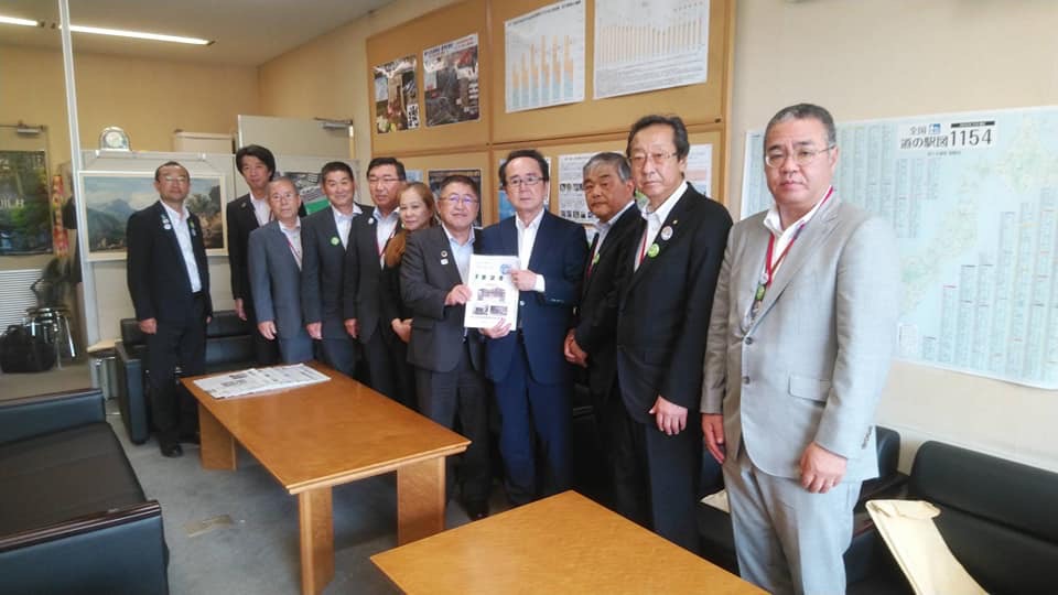 壁にポスターやグラフが飾られた室内で、中央に国土交通省の池田豊人道路局長と市長が一緒に要望書を持ち、9名の関係者が横一列に並んでいる写真