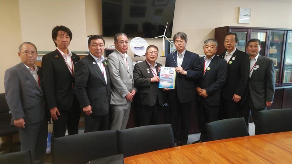 壁上に大画面のモニターが設置された室内で、中央に堀田治官房技術参事官と市長が一緒に要望書を持ち、7名の関係者が横一列に並んでいる写真