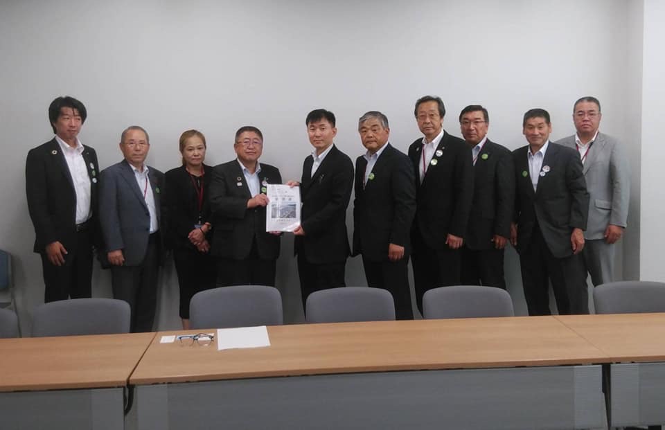 長机が設置された室内で、中央に経済産業省の覚道崇文資源エネルギー政策統括調整官と市長が一緒に要望書を持ち、8名の関係者が横一列に並んでいる写真