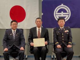 日本国旗、宮古市の市町村旗が掲げられた壁の前で、委嘱状を持つ男性を中心に市長と関係者の男性が椅子に座り横一列に並ぶ記念写真