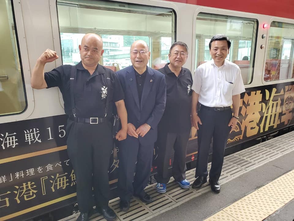 駅のホームで記念ラッピング列車の前で横一列に並ぶ市長と関係者の男性3名を写した記念写真