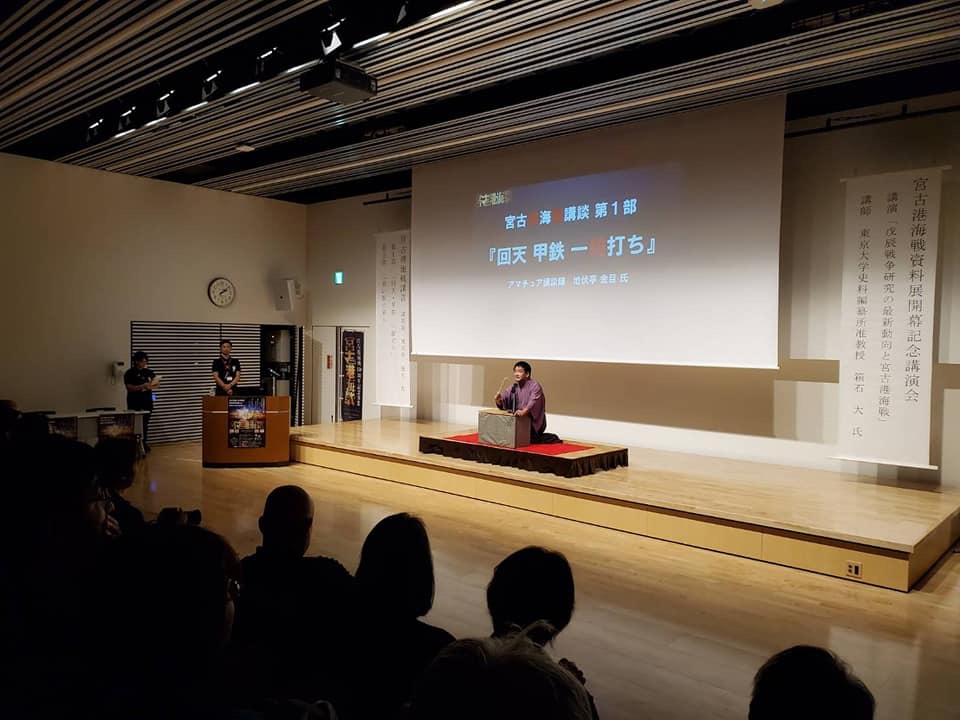 画面が映されたスクリーンが設置された舞台上で講談する様子の地伏亭金目さんの写真