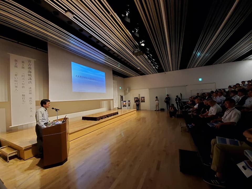 スクリーンの横に置かれた演台に置かれたマイクに向かい講演する箱石大准教授と、階段状に設置された椅子に座り話を聞く様子の参加者たちの写真