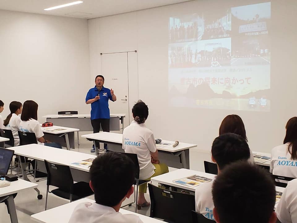画像が投影されたスクリーンの横でマイクを持ち話をする市長と、並べられた机の席に着き話を聞く参加者たちの写真