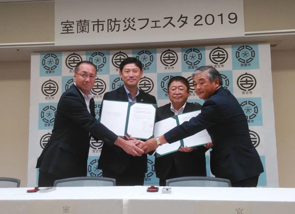 室蘭市と宮古市のバックパネルの前で、協定書を持つ男性と市長、その両側に立つ2名の男性が中央に手を伸ばし握手をしている記念写真