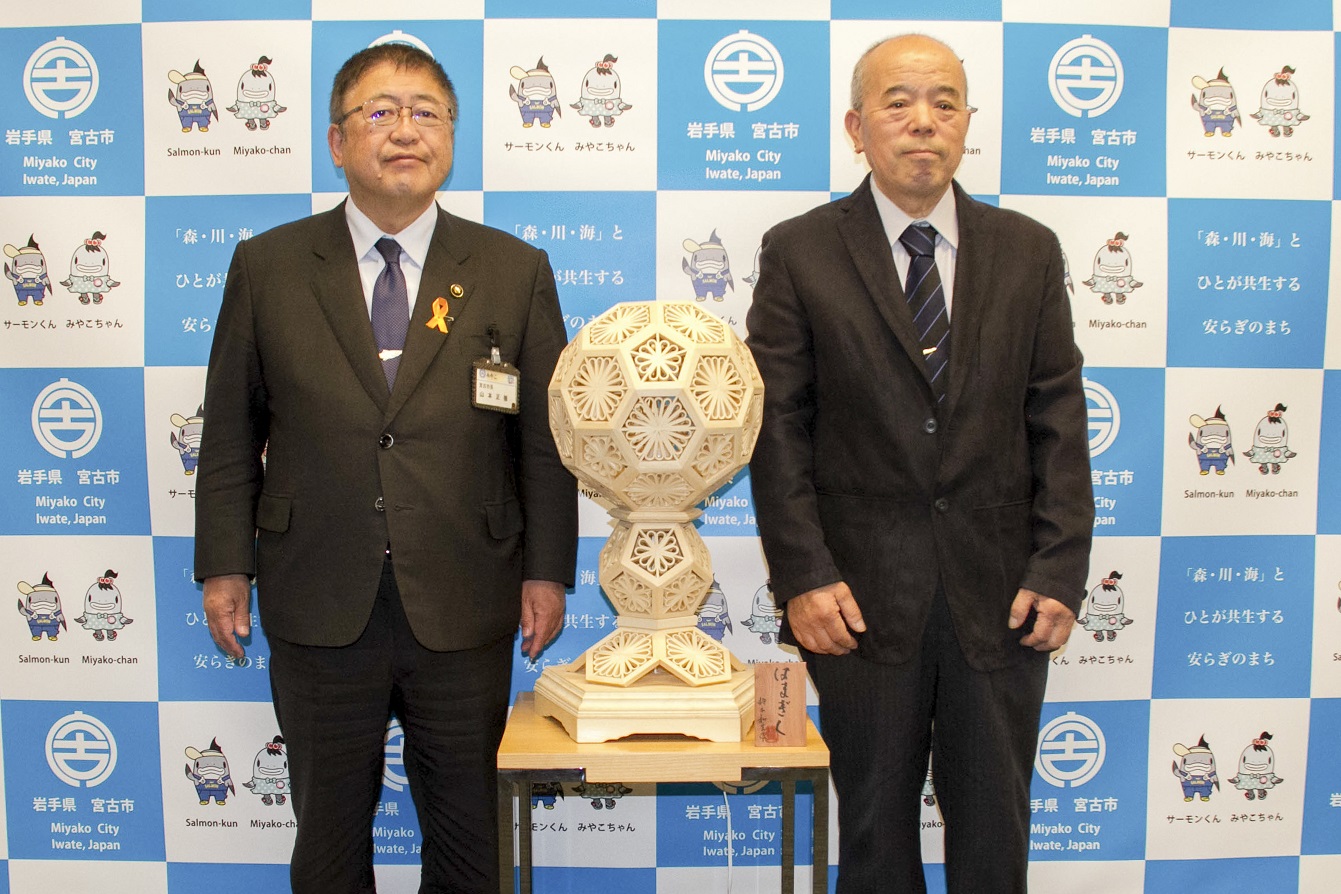 宮古市の花「はまぎく」をモチーフに創作した工芸品の両側に市長と男性が立って並んでいる記念写真