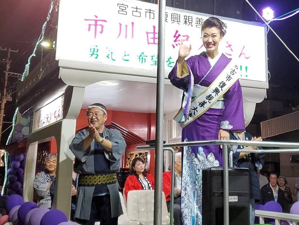 荷台が神輿のようになっている市川由紀乃号に乗り、紫色の着物を着て宮古市復興親善大使のたすきをかけた市川由紀乃さんが手を振り、その後ろで法被を着てハチマキを巻いた市長が手拍子をしている様子の写真