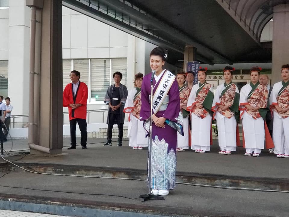 スタンドマイクの前に立ち挨拶をする紫の着物を着た市川由紀乃さんとその後ろに並ぶ白を基調とした和服の衣装を着た女性たちの写真