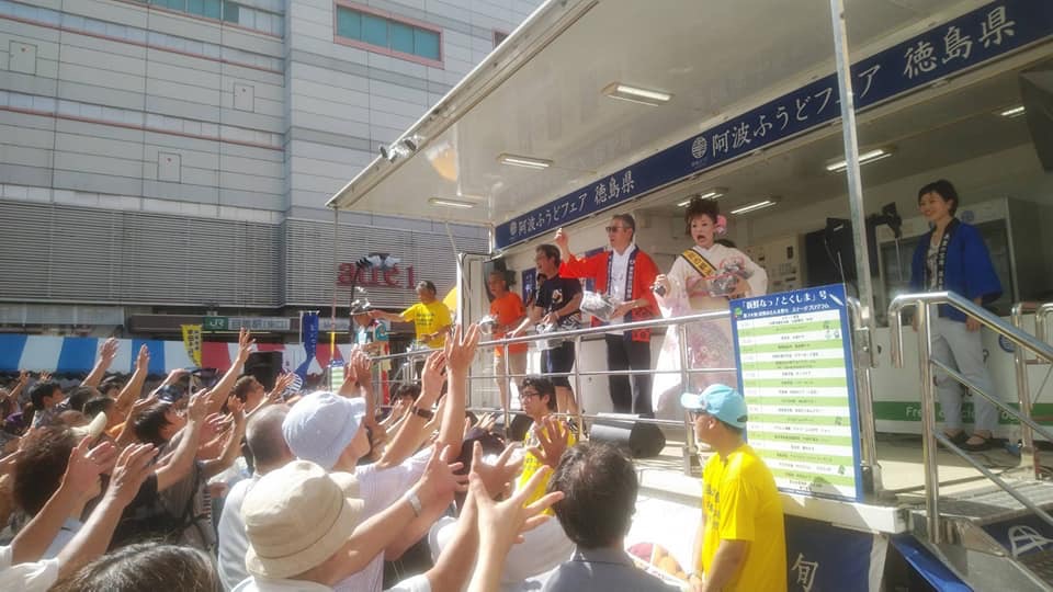 大型ウィング者の荷台から、赤い法被を着た男性や着物の女性が集まったたくさんの来場者の方に向け小さな袋に入った景品を撒いている様子の写真