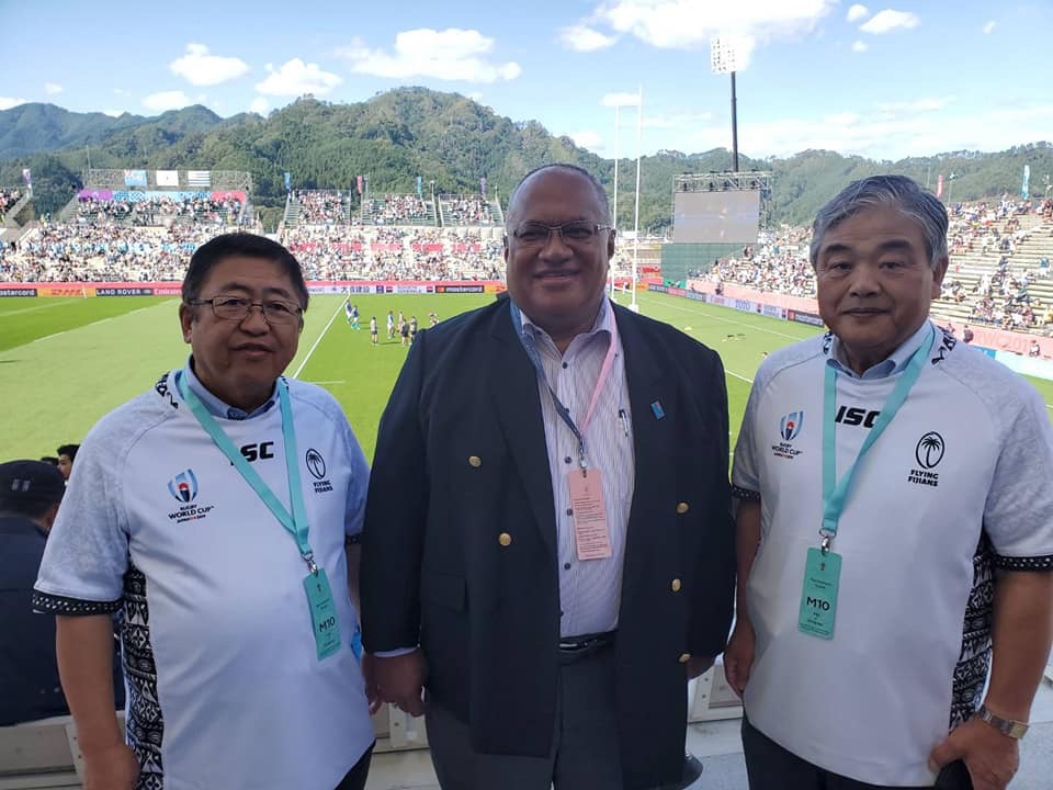 たくさんの観客が押し寄せている晴天の釜石鵜住居復興スタジアムを背景に、市長とイシケリ・マタイトガ全権大使、関係者の男性の3人で撮影された記念写真