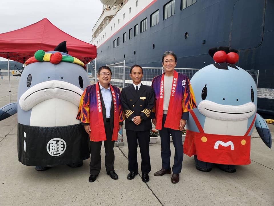 大きなクルーズ船をバックに、法被を着た市長ら2名と制服を着た男性、宮古市のゆるキャラ、サーモンくん・みやこちゃんの集合写真