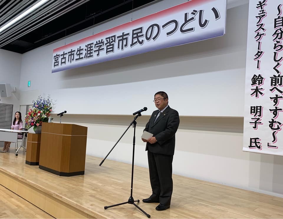 宮古市生涯学習市民のつどいの横断幕が掲げられたステージ上に設置されたスタンドマイクの前に立ち挨拶をする市長の写真