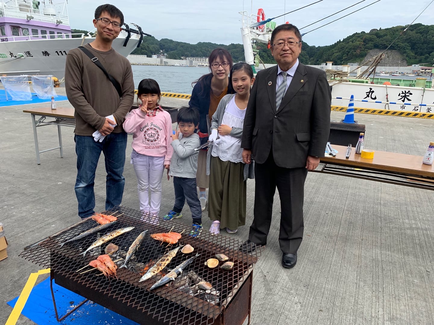 サンマやエビなどの魚介類が焼かれているバーベキューコンロの後ろに立つ、市長と子ども3人を連れた5人家族の写真