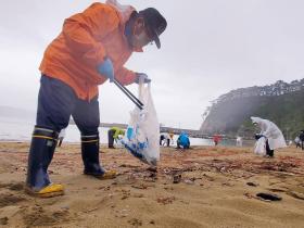 真崎地区の海水浴場で帽子を被り、オレンジ色の作業着を着て、長靴姿の市長が 左手にビニール袋、右手でゴミ拾い用のトングを持ち、清掃活動をしている写真