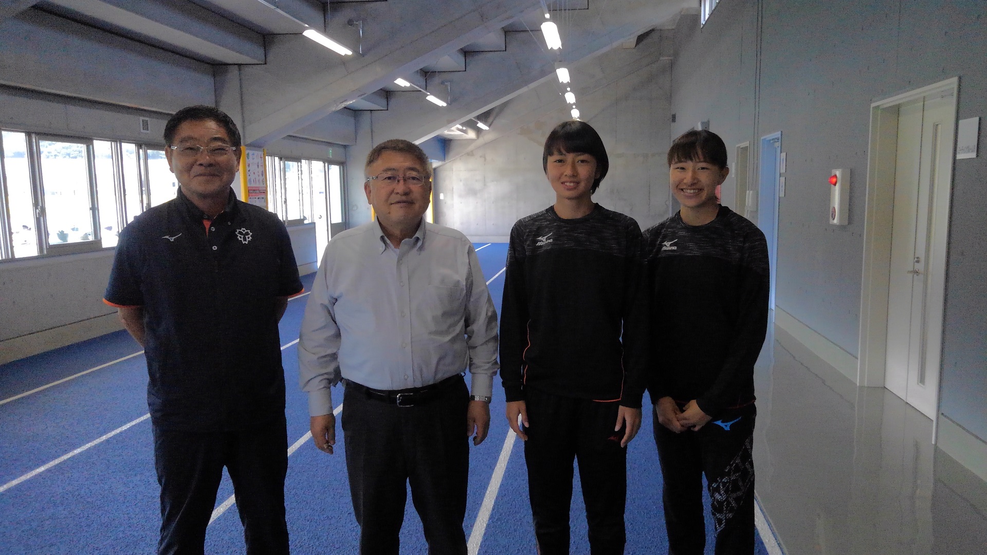青いタータンが敷かれた室内練習場で、市長と日本体育大学陸上競技部 総監督の大村邦英先生、広沢真愛選手、山田未来選手が横一列に並んでいる記念写真